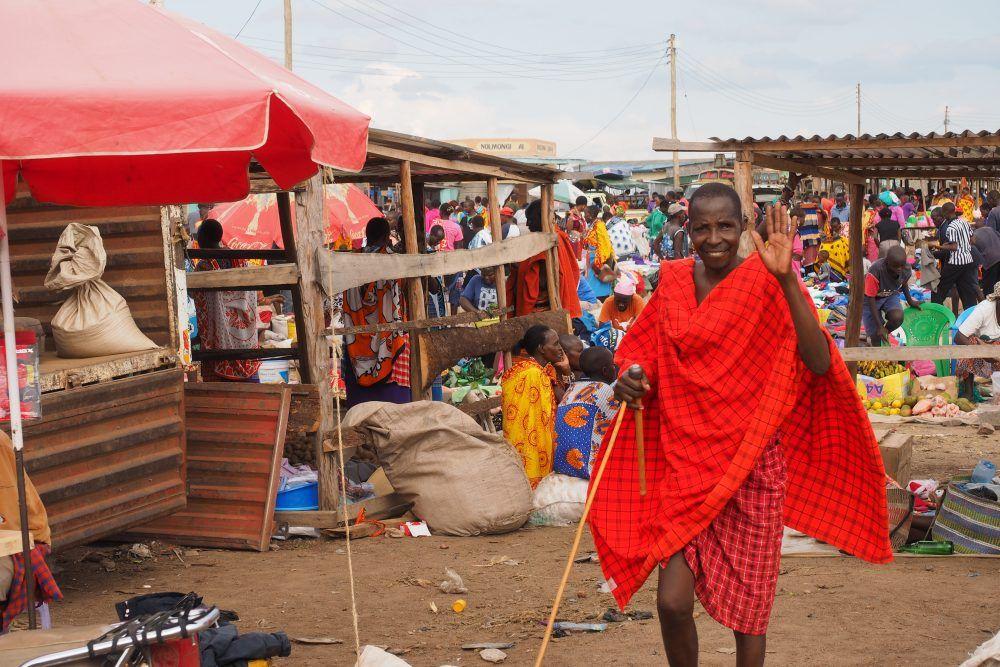 Maasai guide standing in village marketplace waving to camera.