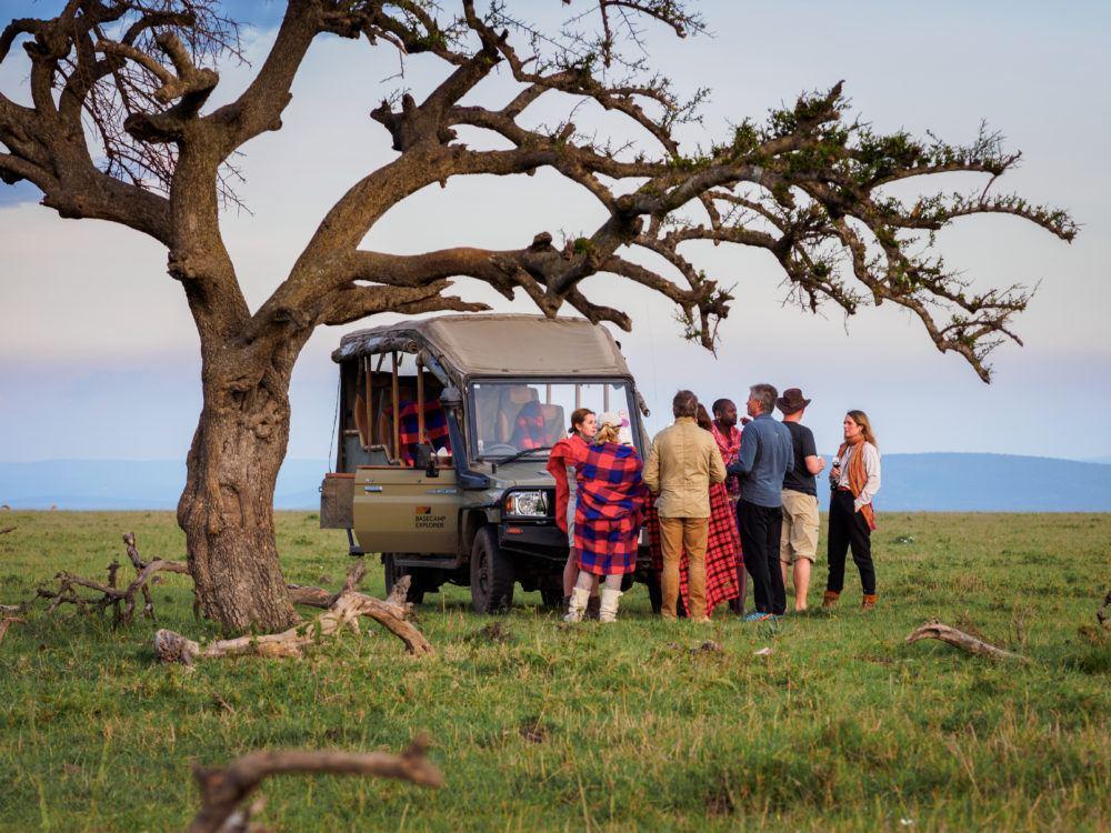 Tourist group on safari game drive standing under tree next to their jeep on the savannah in Masai Mara.